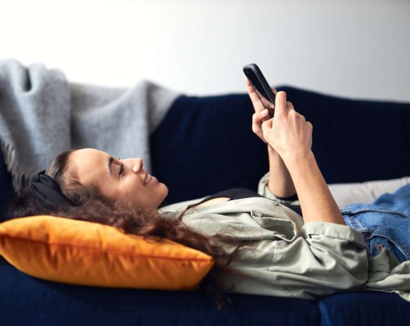 Profile view of young smiling mixed ethnicity woman lying on sofa at home checking social media on mobile phone